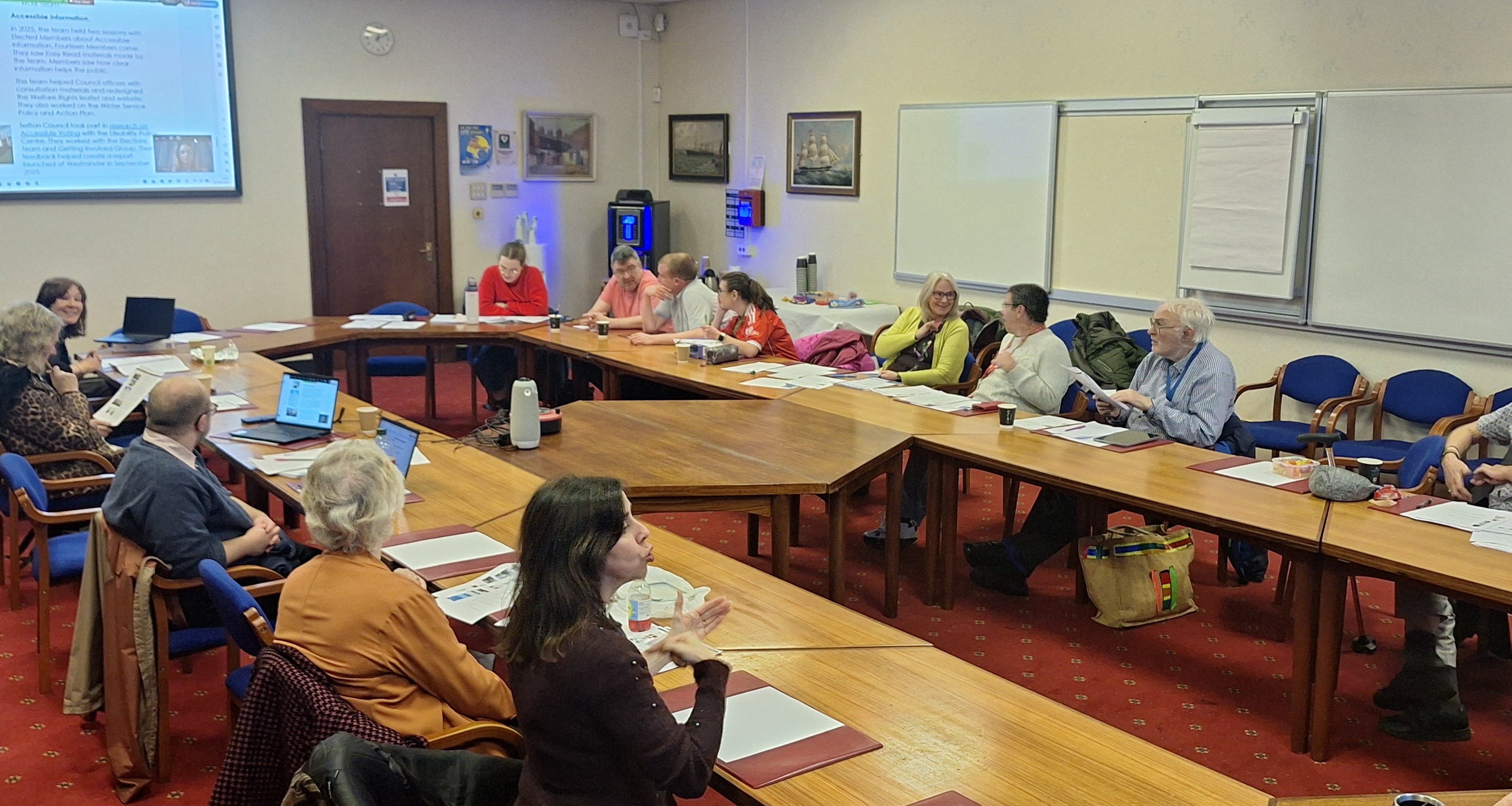 Photograph of the Improving Information Group meeting with member online and in the room, a British Sign Language interpreter is signing for a member of the group. 